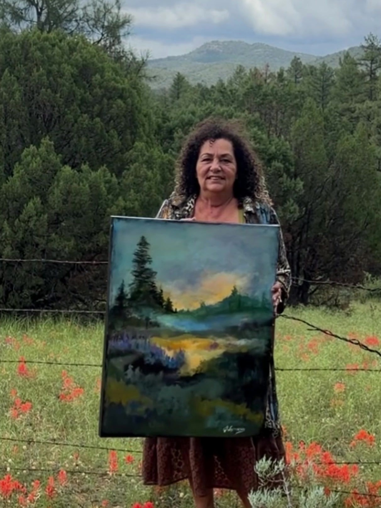 Artist Jeannette Westmoreland holding one of her landscape paintings in a wildflower meadow
